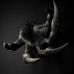 Könnte beinhalten: Eine mattschwarze Skulptur in Form einer Klauenhand mit vier Fingern und einem Daumen, die jeweils in einer scharfen, gekrümmten Kralle enden. Die Oberfläche weist sichtbare Texturlinien und ein dunkles, monochromatisches Finish auf.