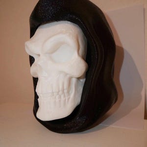Könnte beinhalten: Ein weißer Schädel mit einer schwarzen Kapuze. Der Schädel hat detaillierte Merkmale, einschließlich Augenhöhlen, einer Nasenhöhle und Zähnen. Die Kapuze ist strukturiert und bedeckt die Rückseite und Oberseite des Schädels.