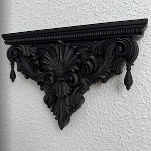 May include: A matte black wall shelf featuring intricate carved scrollwork and acanthus leaf motifs. The shelf has a flat top surface with a beaded edge and two decorative teardrop-shaped finials hanging from the outer edges of the ornate bracket.