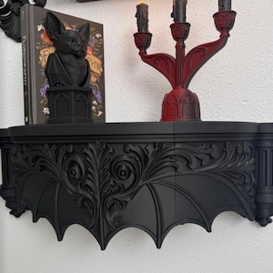 May include: A matte black wall-mounted shelf featuring intricate carved scrollwork and bat-wing shaped edges. The shelf is adorned with detailed foliage patterns and vertical pillar-like supports on each side, creating a structured, symmetrical appearance.
