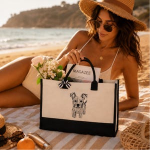 May include: A rectangular canvas tote bag with a black base and black handles. The front features a black line drawing of a small dog and a small black and white striped bow. Inside the bag is a magazine with the text MAGAZEE on the cover.