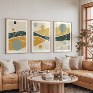 Puede incluir: Tres impresiones artísticas abstractas enmarcadas. Cada impresión presenta un fondo beige con formas geométricas amarillas, verde oscuro y grises. Las impresiones se exhiben sobre un sofá de cuero marrón en un salón.