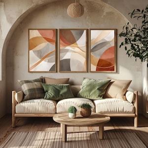 Op de afbeelding: Drie ingelijste abstracte kunstprints met warme tinten oranje, bruin en grijs hangen boven een crèmekleurige bank. De bank is versierd met groene en geruite kussens. Een houten salontafel en vloerkleed maken de woonkamer compleet.