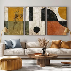 Puede incluir: Tres obras de arte abstractas enmarcadas con formas geométricas y una textura desgastada. La paleta de colores incluye amarillo mostaza, negro, naranja y crema. La obra de arte se exhibe sobre un sofá crema con cojines azules y naranjas.