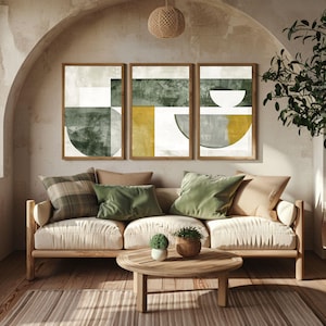 May include: Three framed abstract art prints with geometric shapes in olive green, mustard yellow, and grey tones. The artwork hangs above a cream sofa with green and tan cushions. A round wooden coffee table and a woven rug complete the living room setting.