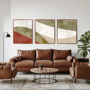 Puede incluir: Tres impresiones artísticas abstractas enmarcadas con un diseño minimalista. Cada impresión presenta formas superpuestas en tonos de rojo, verde, blanco y beige. Las impresiones cuelgan sobre un sofá de cuero marrón y sillas a juego en una sala de estar moderna.