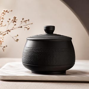Peut inclure: Un pot en céramique noir avec un couvercle. Le pot présente un motif texturé en forme de vagues au milieu et un petit bouton sur le dessus. Il repose sur une surface blanche semblable à du marbre.