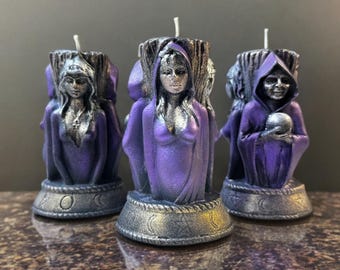 May include: Three purple and silver candles with a mystical theme. Each candle features a sculpted figure in a hooded robe, holding a skull or with a serene expression. The candles have a detailed base with crescent moon designs.