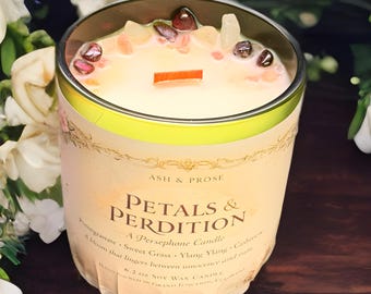 May include: A scented candle in a glass jar, labelled "Petals & Perdition". The candle features a wooden wick and decorative stones. The label lists ingredients including pomegranate and sweet grass. The candle weighs 176 grams.