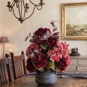 May include: A dark gray, round ceramic vase with two small side handles, filled with a dense arrangement of artificial flowers in deep shades of burgundy, maroon, and muted pink, accented with small red berries and green-tinted foliage.