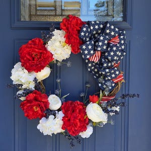 May include: A circular wreath featuring red and white artificial flowers, dark blue sprigs, and a large fabric bow with a white star pattern on a blue background and red and white stripes, all arranged on a natural brown twig base.