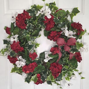 May include: A decorative wreath featuring vibrant red geraniums, white flowers, and lush green ivy. A red and white checkered bow adds a touch of elegance. This floral wreath is perfect for seasonal home decor.
