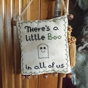 Puede incluir: Decoración cuadrada de Halloween de color blanquecino con el texto "There's a Little Boo in all of us" cosido en negro y verde. Un fantasma sonriente está cosido debajo del texto. Los bordes están adornados con pespuntes verdes y pequeñas campanas.