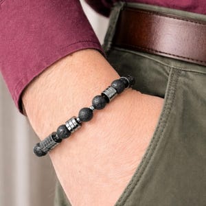 Könnte beinhalten: Schwarzes Perlenarmband mit silberfarbenen Metallakzenten. Das Armband besteht aus runden, porösen schwarzen Perlen und zylindrischen Metallabstandshaltern. Im Hintergrund sind ein weinrotes Hemd und eine olivgrüne Hose zu sehen.
