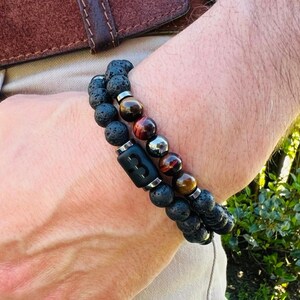 May include: Two beaded bracelets. One bracelet features black lava beads and a black rectangular bead with the number "3". The other bracelet has tiger eye and silver beads. The bracelets are worn on a wrist.