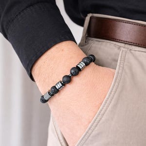 Op de afbeelding: Armband met zwarte lavasteen kralen en zilverkleurige metalen afstandhouders. De armband bestaat uit ronde, poreuze zwarte lavakralen en zilverkleurige metalen cilinders. De armband wordt om de pols gedragen.
