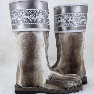 May include: A pair of tall winter boots with a fur-like texture in shades of brown and cream. The boots have a silver-colored cuff with an embroidered floral and reindeer design. The soles are dark brown with white stitching.