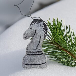May include: A gray felt chess knight ornament with black stitched details. The ornament has a black string for hanging and is set against a snowy background with a pine branch.