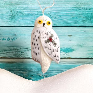 May include: A white felt owl ornament with black spots and yellow eyes. The owl has a small sprig of red berries and green leaves on its wing. A string is attached for hanging. The background is a turquoise wooden surface.