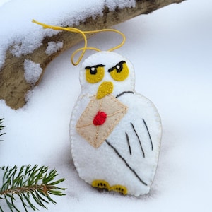 May include: A white felt owl ornament with yellow eyes, beak, and feet, holding a tan envelope with a red heart. The owl has black wing details and a yellow string for hanging. The ornament is set against a snowy background.