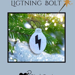 May include: A white felt ornament with a black lightning bolt design. The ornament is embellished with sequins and has a silver cap. The text "LIGHTNING BOLT" is above the ornament. The background features evergreen branches and snow.