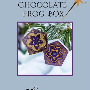 May include: Two felt ornaments in the shape of a pentagon, with a purple base and gold glitter flower design. The ornaments are hanging in front of a snowy background. The text "CHOCOLATE FROG BOX" is at the top.