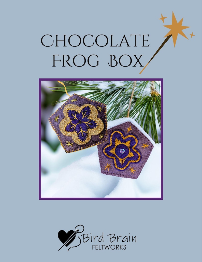 May include: Two felt ornaments in the shape of a pentagon, with a purple base and gold glitter flower design. The ornaments are hanging in front of a snowy background. The text "CHOCOLATE FROG BOX" is at the top.