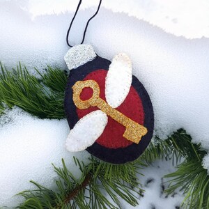 May include: A handmade Christmas ornament shaped like a key with wings. The ornament features a gold glitter key, white wings, and a red circle on a navy blue background. The top is silver glitter.