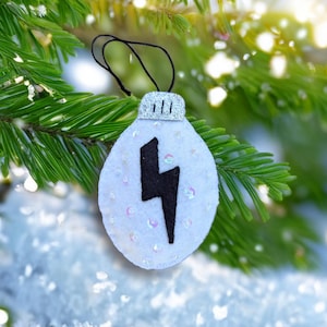 May include: A white felt Christmas ornament shaped like a lightbulb, with a black lightning bolt design. The ornament is decorated with sequins and has a silver glitter top. It hangs from a black string against a backdrop of evergreen branches.