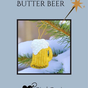 May include: A felt ornament shaped like a mug of butter beer, with a white foamy top and yellow body. The ornament is hanging on a pine branch with snow. The words "BUTTER BEER" are above the ornament.