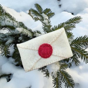 Op de afbeelding: Een handgemaakte, crèmekleurige vilten envelop met een rode, ronde zegel. De envelop is gestikt met een contrasterend draad en ligt op een besneeuwde dennentak. Het ontwerp heeft een vintage, ambachtelijke uitstraling.