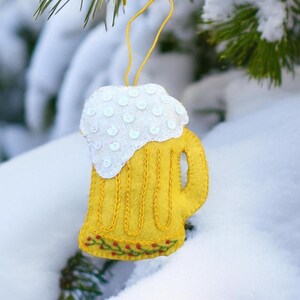 May include: A handmade felt beer mug ornament. The yellow mug has a white foam top embellished with sequins. A yellow string is attached for hanging. The bottom of the mug is decorated with red berries and green leaves.
