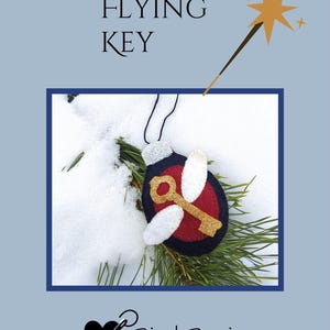 May include: A felt ornament featuring a key with wings, set against a snowy backdrop. The ornament has a red center, gold key, and white wings, with the text "FLYING KEY" above. A gold star wand is also present.