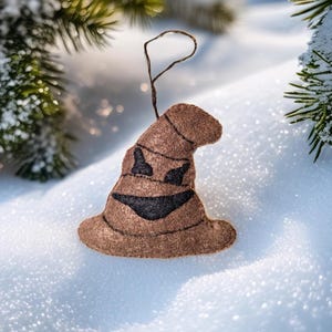 May include: A brown felt Sorting Hat Christmas ornament with a black smiling face. The ornament is hanging from a brown string and is set against a snowy background with evergreen branches.