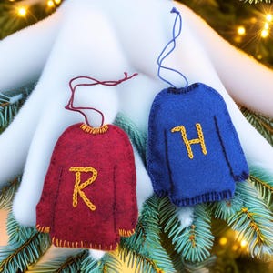May include: Two felt sweater ornaments, one red with a yellow "R" and the other blue with a yellow "H". Both have yellow trim and a string for hanging. They are displayed on a Christmas tree.