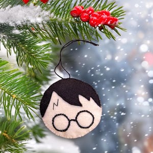 May include: A round, cream-colored Christmas ornament featuring a black-haired character with glasses and a lightning bolt scar. The ornament is hanging from a black string against a blurred background of a Christmas tree with red berries.