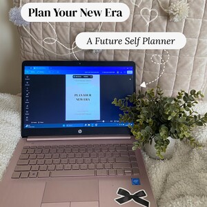 Pode incluir: Um laptop HP rosa dourado exibindo um planejador intitulado "Plan Your New Era" e "A Future Self Planner". Uma pequena planta em vaso está à direita. O laptop está em uma superfície branca com uma almofada decorativa.