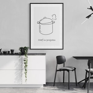 May include: A framed black and white print featuring a line drawing of a cooking pot with steam rising from it. The text "Chef in progress" is below the image. The print is displayed in a modern kitchen setting.