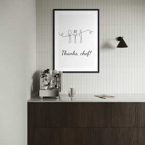 Può includere: Una stampa in bianco e nero incorniciata con la scritta "Thanks, chef!" e un disegno a tratto continuo di un cucchiaio, una forchetta e un coltello. La stampa è sopra un mobile in legno scuro con una macchina per caffè espresso.