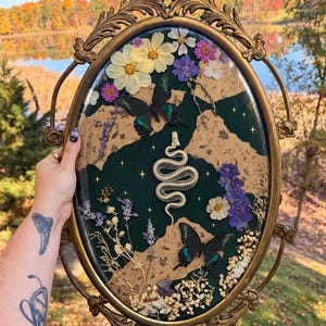May include: An oval wall hanging in an ornate gold frame featuring a central white snake skeleton, two dark green butterflies, and pressed flowers arranged over a background of dark green fabric and torn tan paper with gold star accents.