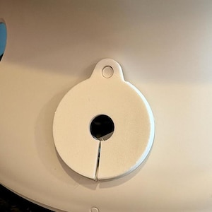 May include: A close-up of a white, circular object with a hole in the center and a slit. The object has a small loop at the top. The background is a light beige color.