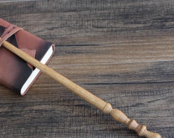 May include: A light brown wooden wand with a smooth, tapered shaft and a handle featuring three rounded, decorative segments. The wand is positioned diagonally across a small, rectangular, brown and black leather-bound notebook.