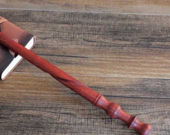 Puede incluir: Una varita mágica de madera marrón rojiza hecha a mano, con un eje en espiral y un mango detallado. La varita está sobre una superficie de madera junto a un cuaderno encuadernado en cuero marrón.
