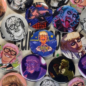 Puede incluir: Una colección de botones redondos con varias ilustraciones. Los botones presentan retratos de una persona con gafas, auriculares y otros diseños gráficos. Algunos botones tienen texto, y la paleta de colores incluye tonos de azul, rosa y negro.