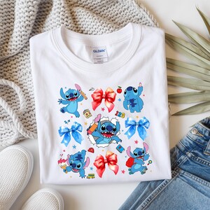 Op de afbeelding: Een wit T-shirt met korte mouwen en een kleurrijke grafische print van het blauwe personage Stitch in verschillende houdingen, afgewisseld met rode en blauwe strikken, kleine schoolgerelateerde iconen en een centrale afbeelding van Stitch die uit gescheurd papier komt.