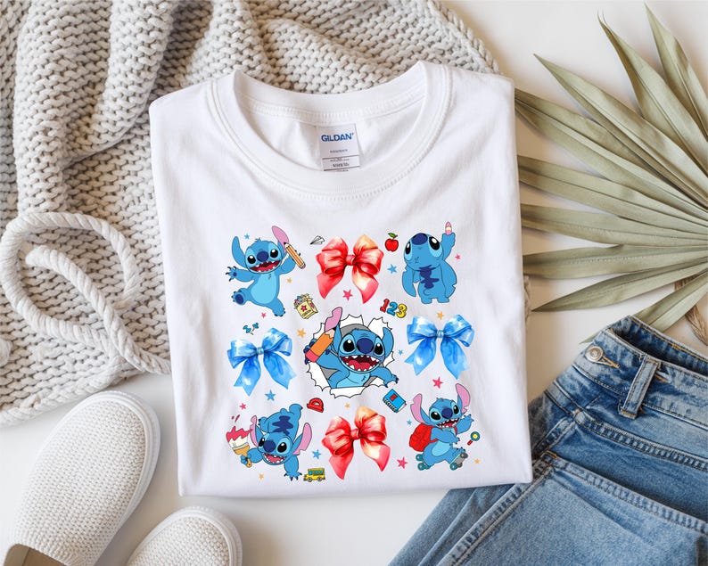 Op de afbeelding: Een wit T-shirt met korte mouwen en een kleurrijke grafische print van het blauwe personage Stitch in verschillende houdingen, afgewisseld met rode en blauwe strikken, kleine schoolgerelateerde iconen en een centrale afbeelding van Stitch die uit gescheurd papier komt.