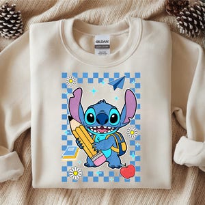 Może przedstawiać: Kremowa bluza z okrągłym dekoltem z grafiką przedstawiającą niebieską postać Stitcha trzymającą duży ż&oacute;łty oł&oacute;wek. Wz&oacute;r zawiera niebieskie tło w kratkę, czerwone jabłko, papierowy samolot, stokrotki oraz małe niebieskie ikony gwiazdek.