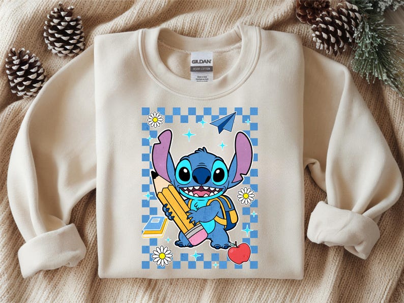 Może przedstawiać: Kremowa bluza z okrągłym dekoltem z grafiką przedstawiającą niebieską postać Stitcha trzymającą duży ż&oacute;łty oł&oacute;wek. Wz&oacute;r zawiera niebieskie tło w kratkę, czerwone jabłko, papierowy samolot, stokrotki oraz małe niebieskie ikony gwiazdek.