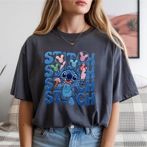 May include: Dark gray t-shirt featuring a blue cartoon character holding ice cream cones. The word "STITCH" is printed in blue, with the character's name repeated multiple times. The shirt is a relaxed fit.