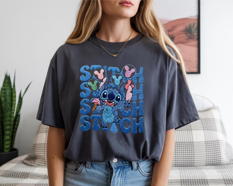 May include: Dark gray t-shirt featuring a blue cartoon character holding ice cream cones. The word "STITCH" is printed in blue, with the character's name repeated multiple times. The shirt is a relaxed fit.